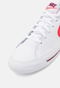 Zapatilla Nike blanca con cordones blancos, swoosh rojo y logo de Nike rojo en la lengüeta, mostrada desde un ángulo cercano frontal.