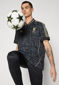 Fekete mintás mez arany részletekkel, amelyen Liverpool FC logó látható, fekete sportnadrággal párosítva. Egy focilabda a modell térde közelében lebeg.