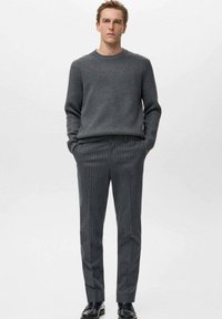 Maglione grigio a costine con scollo rotondo, abbinato a pantaloni grigi a righe sottili. Il modello sta in piedi con sicurezza con le mani nelle tasche.