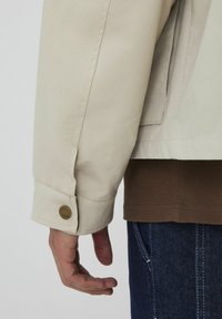 Veste beige avec un poignet boutonné, en tissu texturé, superposée sur une chemise marron à côtes, associée avec un jean en denim bleu.