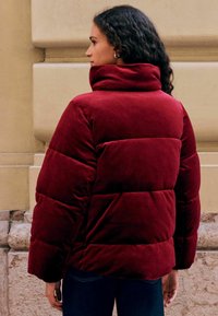 Burgundy puffer jacket with a high collar, featuring quilted sections and a soft, velvety texture. Styled with dark blue jeans.