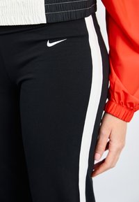 Čierne športové nohavice s bielymi bočnými pruhmi, s bielym logom Nike na stehne. Hladká textúra; kombinované s červenou bundou.
