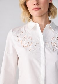 Camicia bianca di cotone con ricami floreali sul petto e dettagli a taglio, con colletto a bottoni e maniche lunghe.