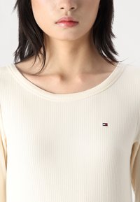 Rippetop mit langen Ärmeln in Creme mit weitem Ausschnitt und kleinem gesticktem Logo in Rot, Weiß und Blau auf der Brust.