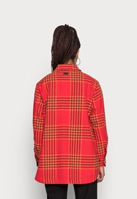 Rød- og orangeternet skjorte med knap-ned krave, tekstureret stof, lange ærmer og et diskret sort mærke bagpå.