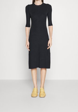 Femme portant une robe midi noire côtelée avec des épaules bouffantes et des manches jusqu'au coude, associée à des chaussures jaunes à enfiler.