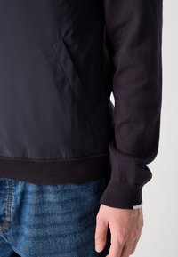 Σκοτεινός μπουφάν με απαλό υφασμάτινο πάνω μέρος και πλεκτούς μανσέτες; διαθέτει πλευρική τσέπη και ριμπ στα τελειώματα; φορεμένος πάνω από μπλε τζιν παντελόνι.