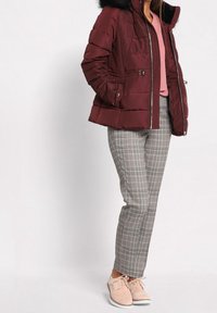 Veste matelassée bordeaux avec capuche en fourrure, fermeture éclair sur le devant, deux poches latérales ; associée à un pantalon à carreaux gris et des chaussures roses.