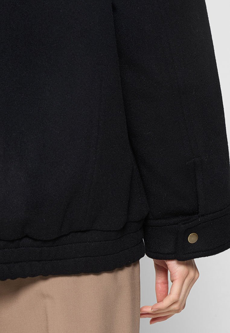 Schwarze Wollmischungjacke mit gewalzten Textur, ausgestattet mit einem taillierten Saum, sichtbaren Nähten und einem Druckknopfdetail in goldfarbener Ausführung.