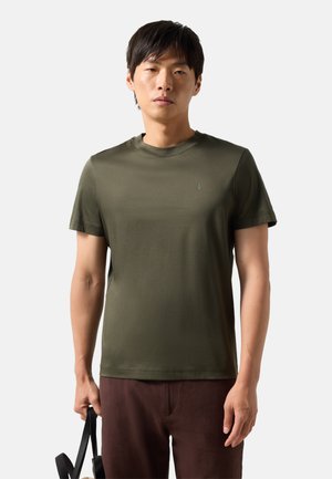 Mężczyzna ubrany w oliwkowozieloną koszulkę z krótkim rękawem i brązowe spodnie, trzymający torebkę z czarnymi rączkami na jednolitym jasnym tle.