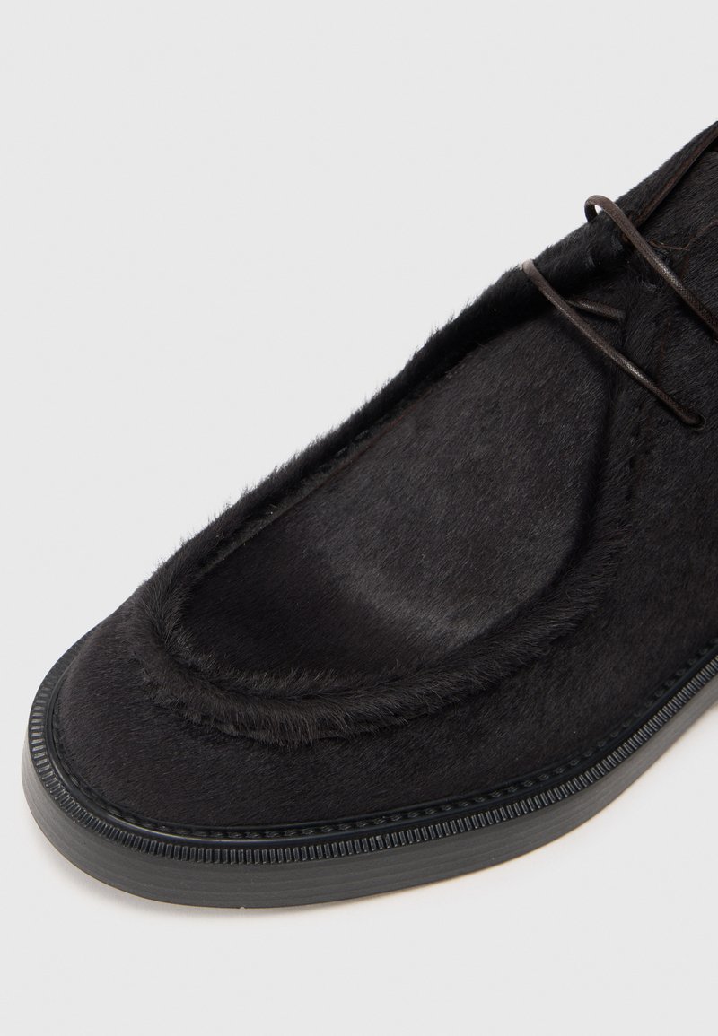 Sort sko med blødt, tekstureret overdel, der ligner hår, rund tå, syet detaljer og kontrasterende gummisål. Snørebåndene er sikret ovenpå.