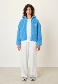 Blauwe fleece jack met rits van een textuur met pluizig materiaal, gecombineerd met grijze sweatpants en grijze sneakers. Witte onderhemd zichtbaar.