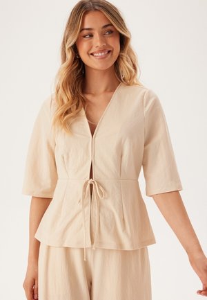 Beige peplum top med korte ærmer, V-udskærkning og snorelukning i taljen. Blødt struktureret stof med en afslappet pasform.