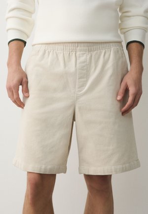 Mand iført beige kordfløjls knælange shorts og cremefarvet langærmet trøje, stående med afslappede hænder langs siderne.