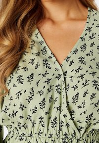Robe fleurie verte avec un décolleté en V, motifs de feuilles noires et une taille élastique pour une silhouette ajustée. Tissu lisse avec une texture légèrement brillante.