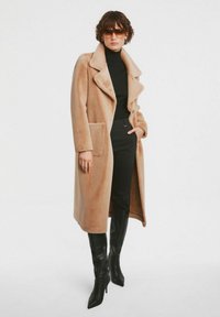 Beżowy długi płaszcz wełniany z kołnierzem typu klapa, zapięciem na guziki i przednią kieszenią. Nosi się go na czarną bluzkę, w komplecie z czarnymi wysokimi botkami do kolan.