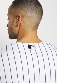 Fehér póló fekete függőleges csíkokkal, textúrált anyagból és kerek nyakkivágással. A gallérnál a Major League Baseball logós címke látható.