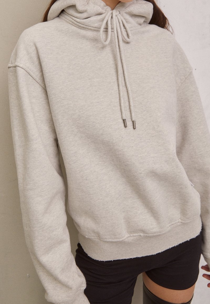 Světlá šedá mikina s kapucí na šňůrku, žebrovanými manžetami a surovým lemem. Vyrobena z měkké látky, má volný střih a minimální detaily.
