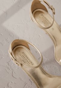 Paire de sandales métalliques dorées à bout ouvert avec courroies à la cheville, semelles plates, et marque "Signature Made in Brazil" à l'intérieur.