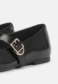 Sorte patentlærersko med flad sål og ankelrem med gyldent spænde. Glat, skinnende finish og strømlinet design.