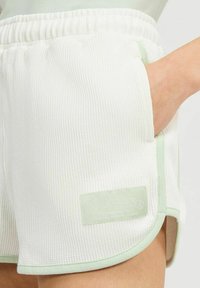 Pantalones cortos blancos con textura y detalles en verde claro, con un bolsillo frontal, llevados por una persona con la mano dentro del bolsillo.