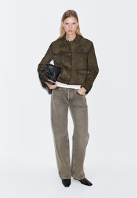 Giacca in suede verde scuro, design cropped con chiusura a bottone. Abbinata a jeans beige sbiaditi a gamba larga e scarpe nere a punta. Tiene una borsa nera.