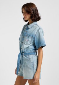 Ljusblå denimshortsdräkt med korta ärmar, framfickor och matchande bälte. Har en blekt tvätt och råa fållar.