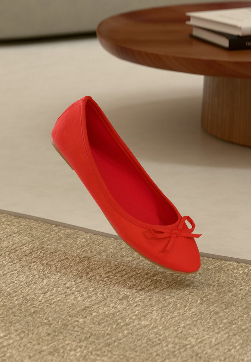Red flat shoe featuring a rounded toe, a small bow detail on the front, and a smooth textile upper, set against a textured surface.