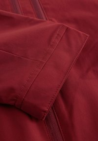 Détail d'une veste en tissu rouge présentant une texture lisse, des coutures visibles et une fermeture éclair dissimulée sur un côté.