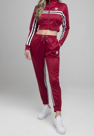 Vrouw die een rode, korte ritsvest en bijpassende joggingbroek met witte en zwarte strepen draagt, gecombineerd met witte platformsneakers.