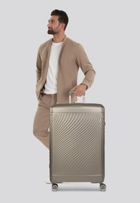 Gray hard-shell suitcase with a wave pattern, metal handle, and four rolling wheels. Smooth texture and sleek design.