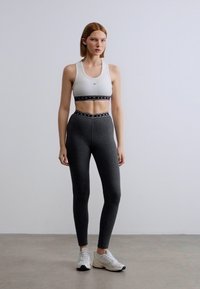 Λευκό αθλητικό σουτιέν με μαύρο περίγραμμα· σκούρο γκρι, εφαρμοστά κολάν με σχεδιασμένη ζώνη μέσης· σετ με ελαφριά αθλητικά παπούτσια.