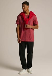 Czerwona koszulka z teksturą w połączeniu z czarnymi spodniami, białymi sneakersami i czerwonym swetrem zarzuconym na ramiona. Prezentacja casualowego stroju.