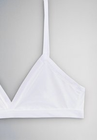 Biely trojuholníkový bralette s tenkými ramienkami a bez viditeľných zapínaní, zobrazený na jednofarebnom sivom pozadí.