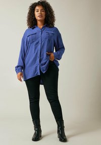 Fioletowa koszula z guzikami, z dwoma kieszeniami na piersiach, luźny krój i zaokrąglony dół, zestawiona z czarnymi skinny jeansami i czarnymi botkami za kostkę.