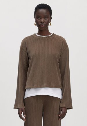 Woman wearing brown ribbed long-sleeve top and matching pants with white shirt visible underneath, gold hoop earrings, neutral background.