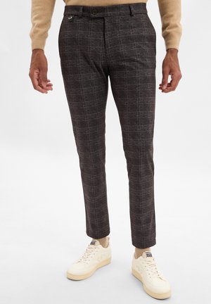 Dunkelkarierte Hosen aus einem strukturierten Stoff, mit einer Knopfverschlus und Seitentaschen, kombiniert mit cremefarbenen Sneakers und einem beigen Pullover.