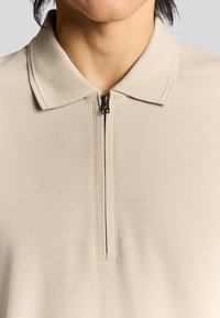 Beige zip-polo tröja i ett texturerat tyg, med en klassisk krage och en kort dragkedja vid halsen.