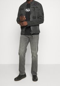 Harmaa farkkutakki, jossa on teksturoituja applikaatioita ja vetoketjusuljenta, yhdistettynä mustaan t-paitaan, harmaihin farkkuihin ja mustiin kenkiin, esitetty valkoisen taustan edessä.