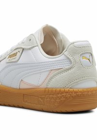 Fehér sneaker sima bőr felsőrésszel, világosszürke nubukbőr kiegészítésekkel és texturált gumitalppal. Arany PUMA logó a saroknál.