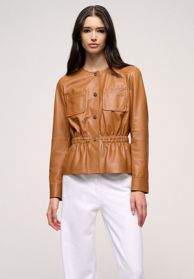 Brown leather jacket with a cinched waist, snap buttons, and two chest pockets. Styled with white pants for contrast. Smooth texture.