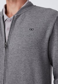Felpa grigia con zip e un motivo testurizzato, dotata di colletto rialzato e un piccolo logo nero sul petto. Collo con bottoni sotto.