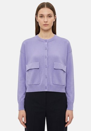 Donna che indossa un cardigan lavanda con bottoni e due tasche con pattina sul davanti, abbinato a pantaloni neri, in piedi su uno sfondo bianco.