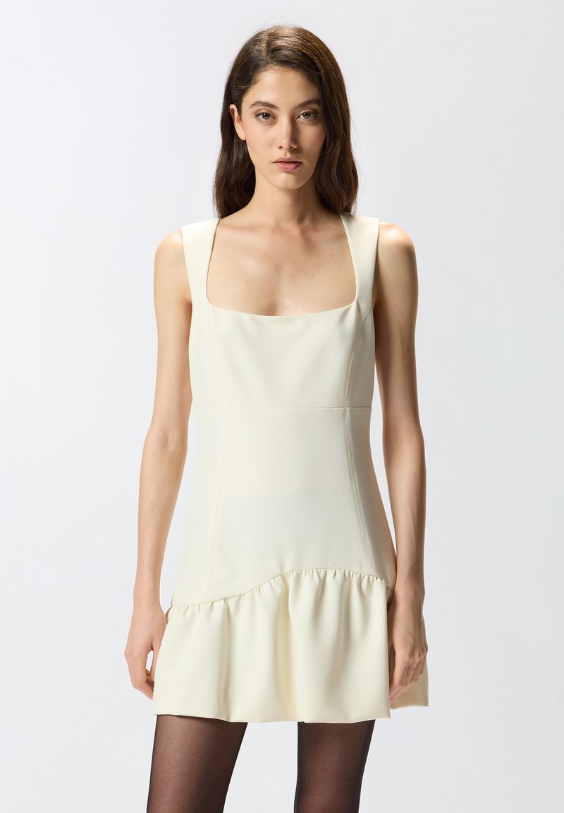 Vestido sin mangas de color crema con escote cuadrado, cuerpo ajustado y dobladillo acampanado con volantes; textura suave, diseño minimalista.