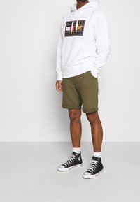 Hvid hættejakke med blåt og rødt grafisk emblem, parret med olivengrønne shorts og sorte højtoppede sneakers med hvide detaljer.