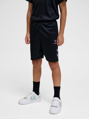 Dziecko ubrane w czarne krótkie spodenki sportowe z fioletowym logo i strzałkami, czarne skarpetki i białe trampki z niebieskimi szewronami, stojące na jednolitym tle.