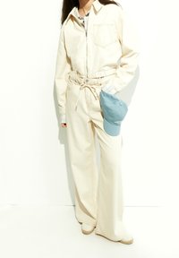 Krämfärgad jumpsuit med dragkedja, elastisk midja och framficka. Parat med blå keps och vävda tofflor. Texturerad tyg.