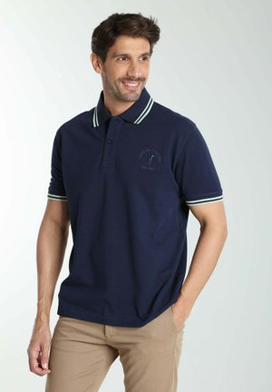 Homme souriant portant un polo bleu marine avec des rayures blanches sur le col et les manches, et un pantalon beige, debout avec une main dans la poche.