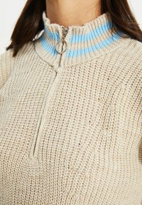 Beige gestrickter Pullover mit einem Viertelreißverschluss, ausgestattet mit einem gerippten, gestreiften Kragen in Blau und Creme sowie strukturiertem Stoff. Sichtbare gestickte Details.