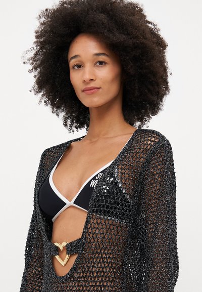 Cubierta de ganchillo negra con tejido abierto, mangas largas y un acento dorado en forma de corazón en la cintura, superpuesta sobre un bikini negro.